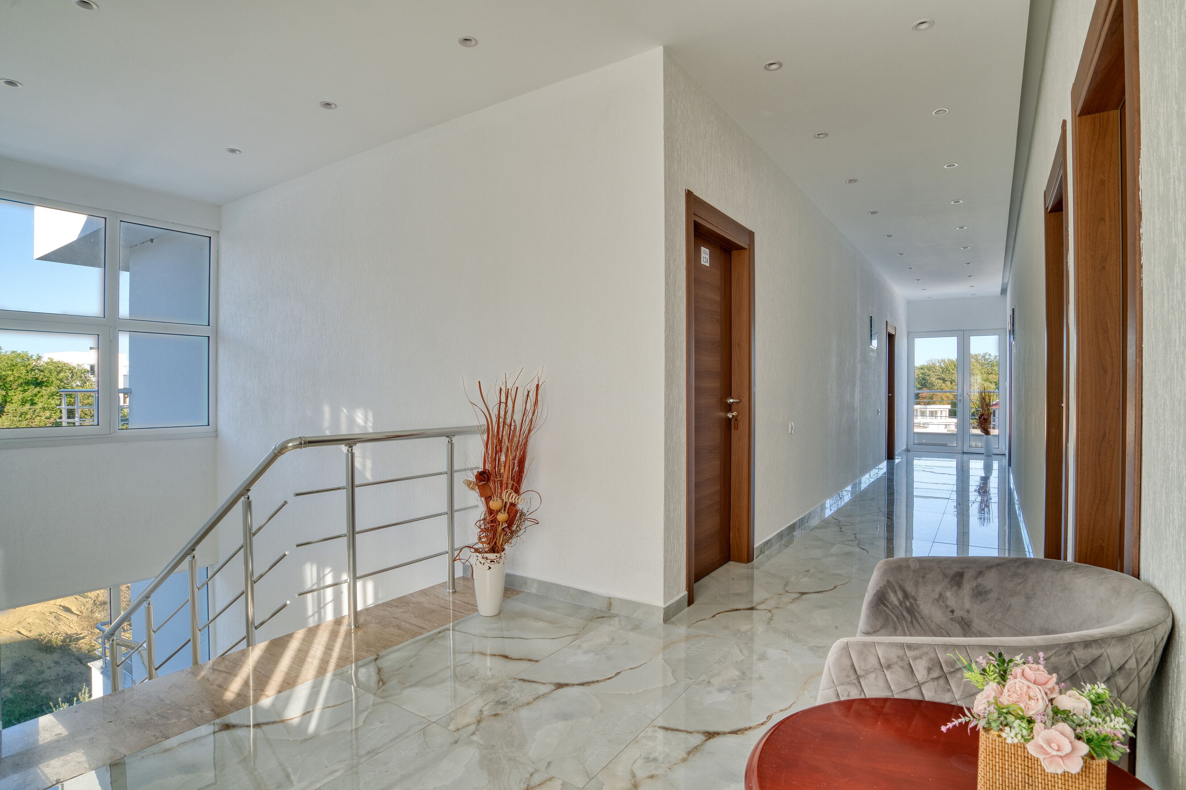 A modern hotel lobby with a marble floor, a staircase, a wooden door, and a seating area with a vase of flowers.