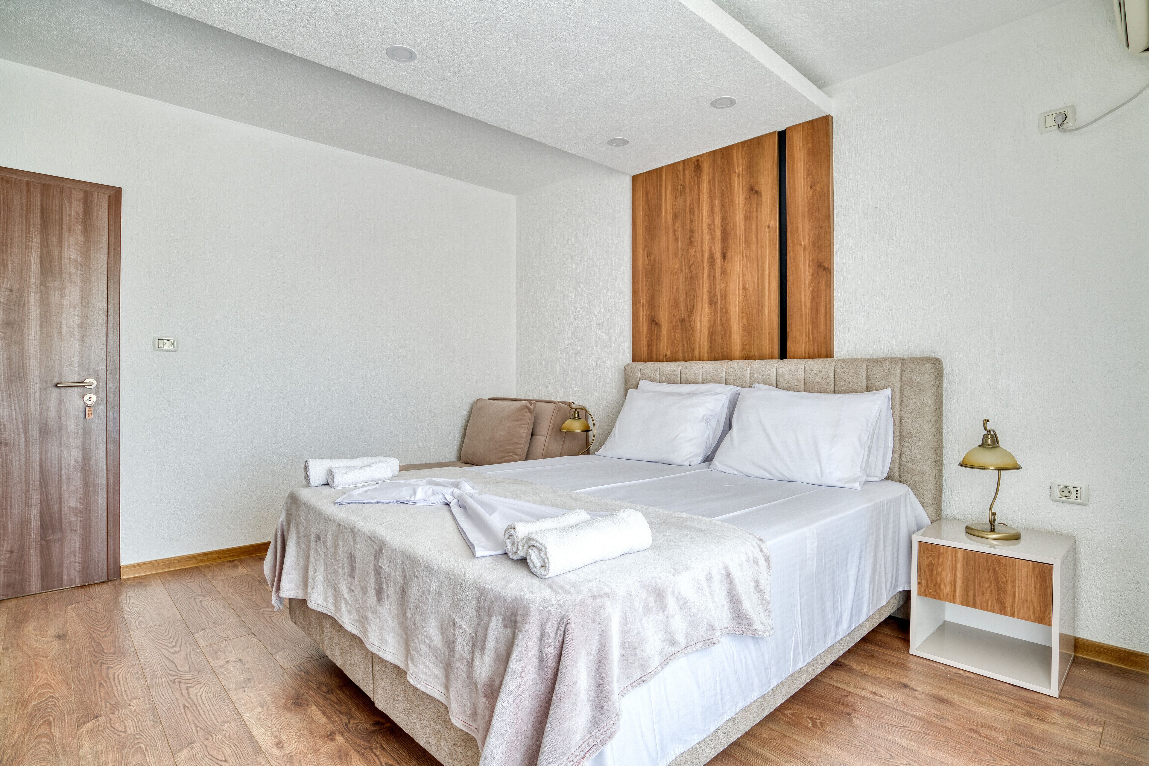 A bedroom with a wooden floor, a bed with white linens, a bedside table with a lamp, and a wooden wardrobe.