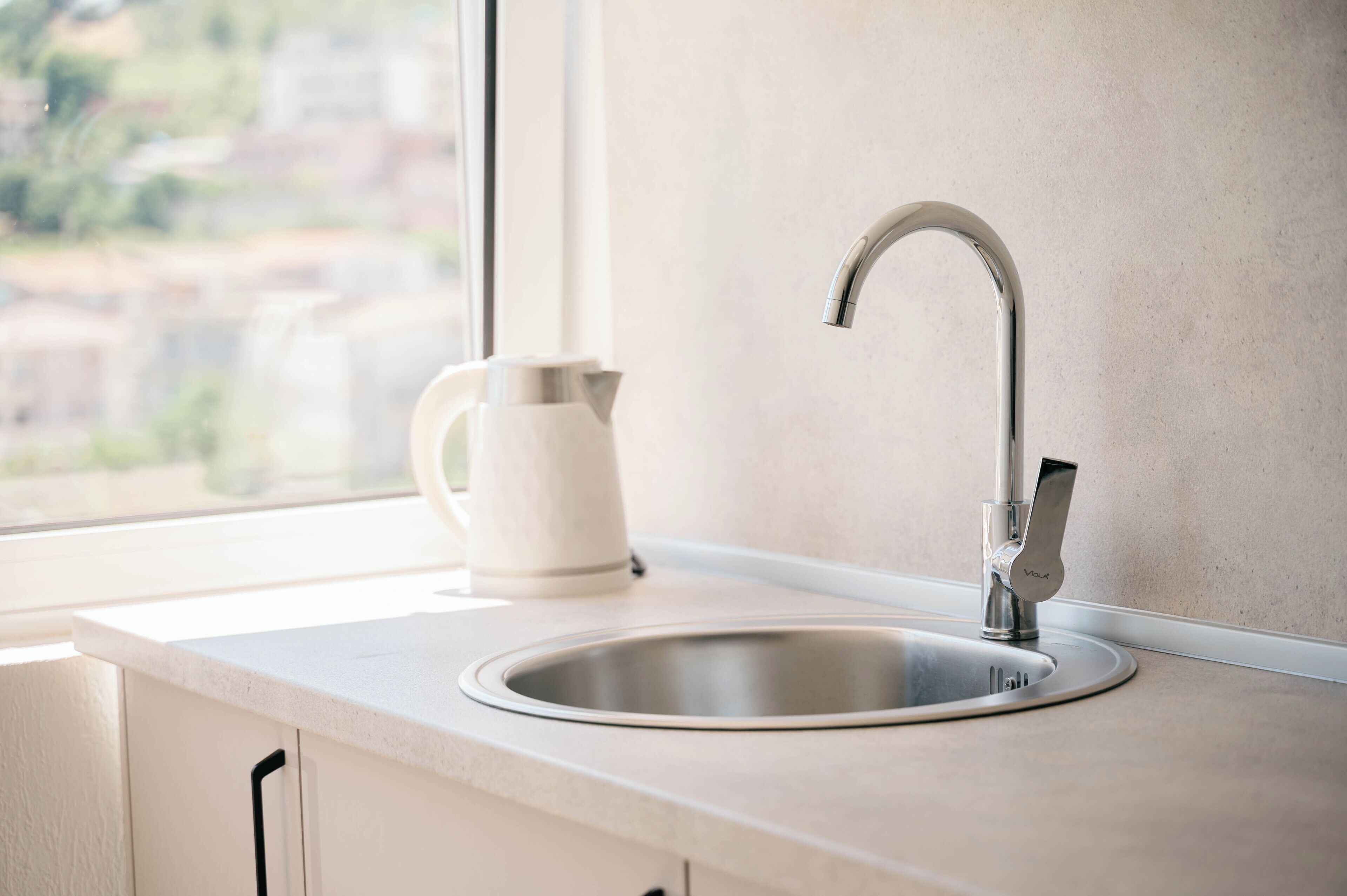 A kitchen with a stainless steel sink, a kettle, and a window providing a view of the outside.