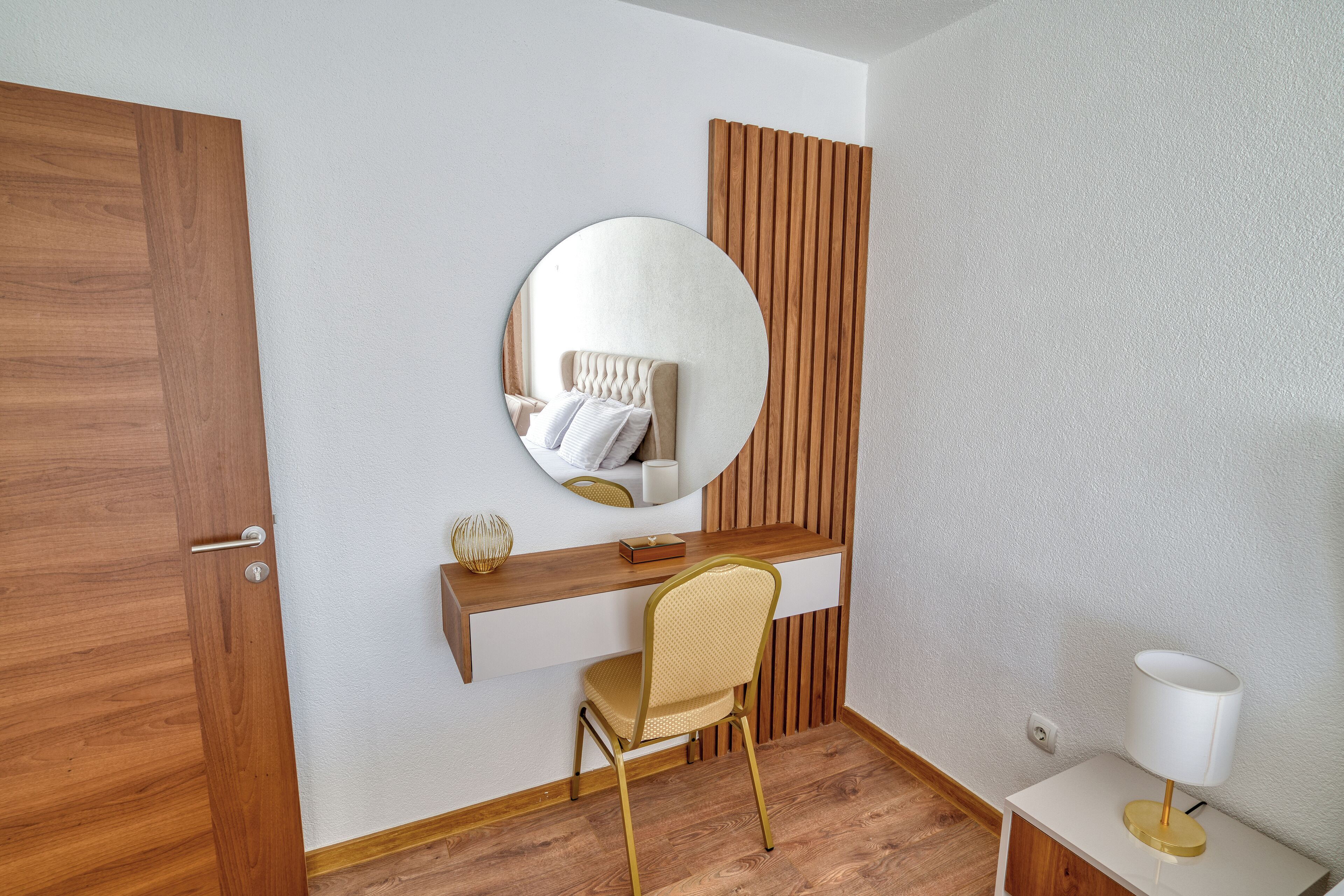 A modern room with a wooden door, a round mirror, a white desk, a chair, and a bedside table with a lamp.