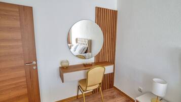 A modern room with a wooden door, a round mirror, a white desk, a chair, and a bedside table with a lamp.