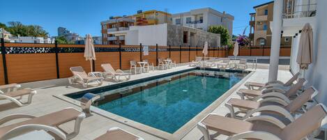 A poolside area with lounge chairs and umbrellas, surrounded by a wooden fence and buildings in the background.