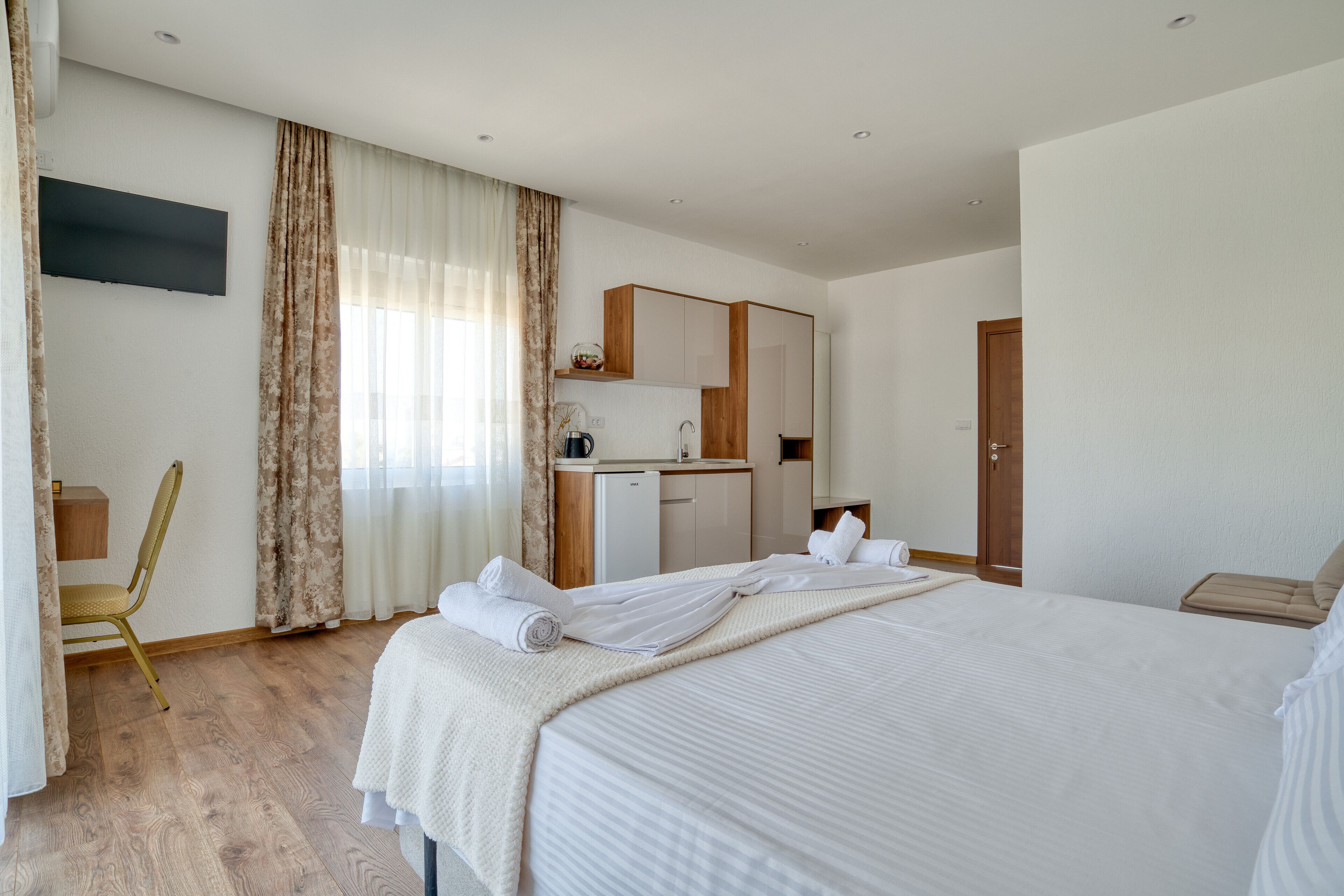 A modern hotel room with a large bed, a desk, a kitchenette, and a bathroom.