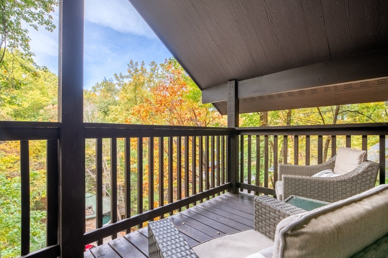 Une véranda couverte avec une balustrade en bois, des meubles en osier et une vue sur le feuillage automnal.