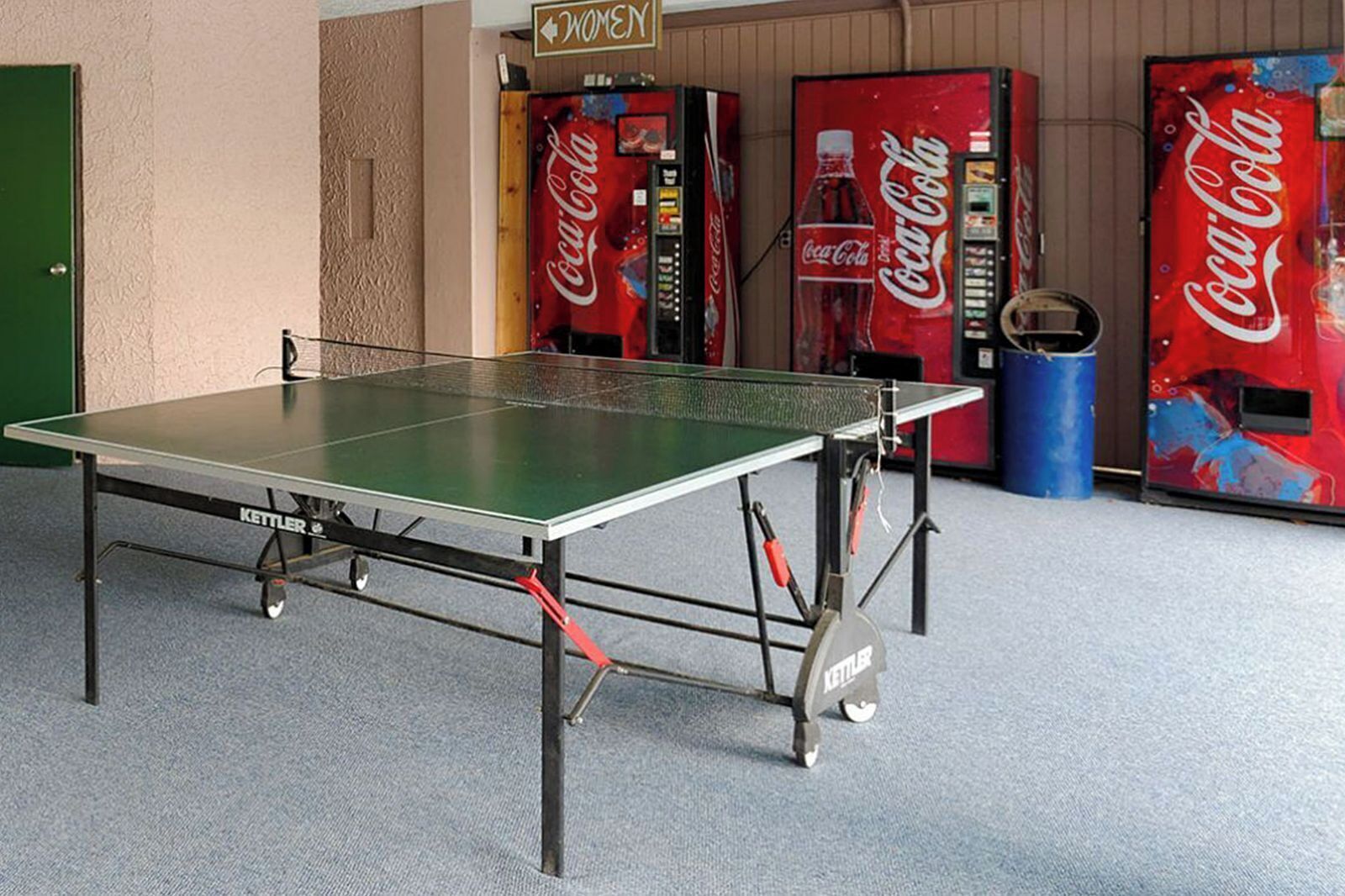 A ping pong table in a room with vending machines, including Coca-Cola and a sign for "WOMEN".