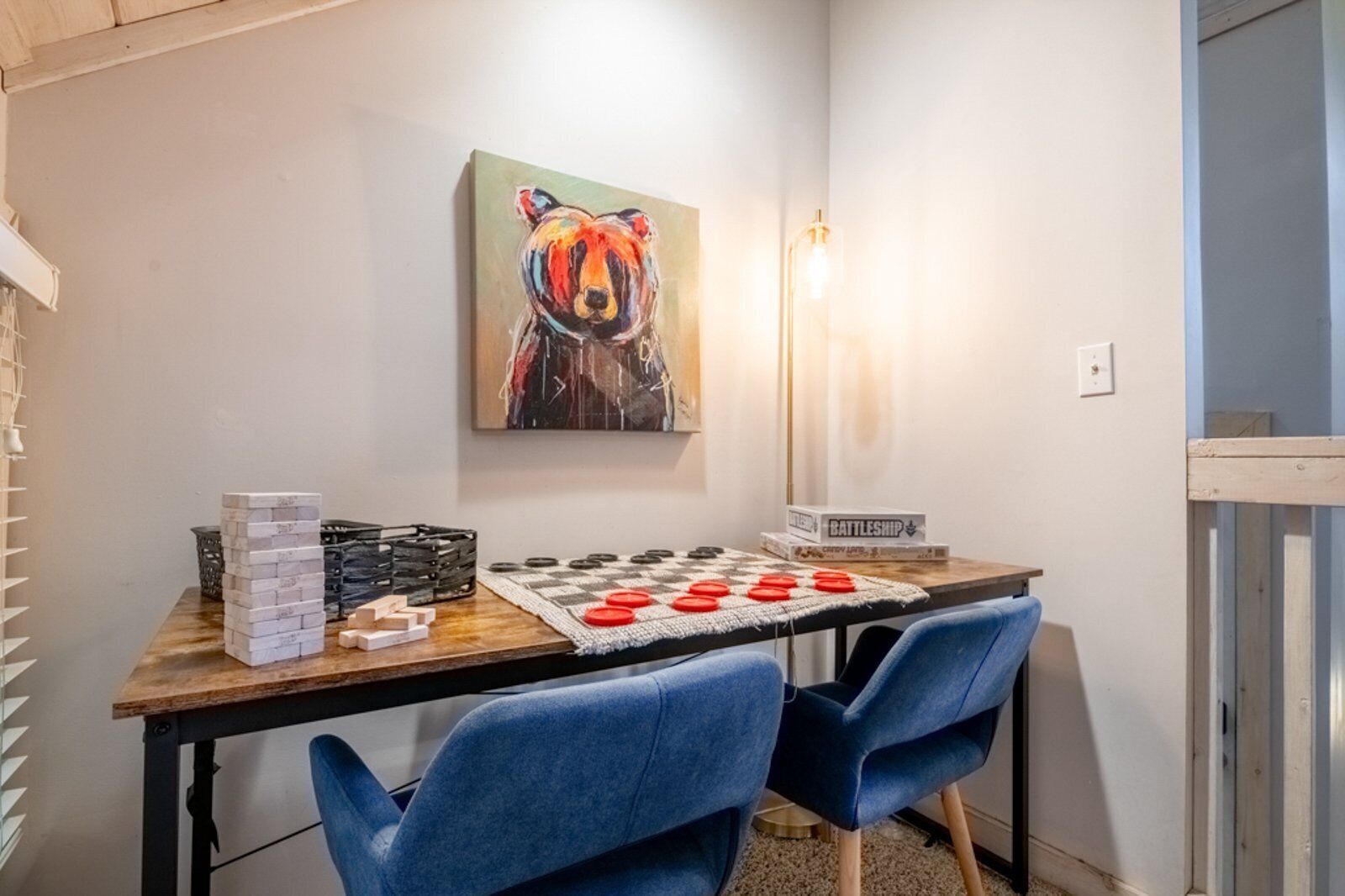 A room with a wooden table, two blue chairs, a board game, a Battleship game, and a colorful bear painting on the wall.
