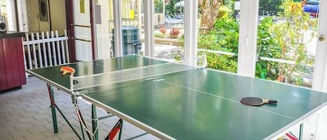 Ein Tischtennis-Tisch mit Schlägern, aufgestellt in einem Raum mit großen Fenstern und Blick ins Freie.