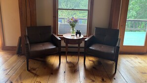 A room with two leather chairs, a small table with a vase of flowers, and a large window with a view of greenery. - Tranquil lodge Ski. Stay. Hike. Soak in the River and Views. (Brookline)