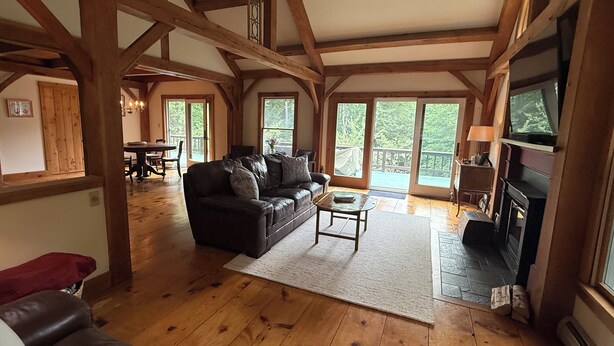 A spacious living room with a wooden ceiling, a fireplace, and a large window overlooking a deck. - Tranquil lodge Ski. Stay. Hike. Soak in the River and Views. (Brookline)