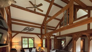 A spacious living room with a high ceiling, wooden beams, a fireplace, leather sofas, and a large window. - Tranquil lodge Ski. Stay. Hike. Soak in the River and Views. (Brookline)
