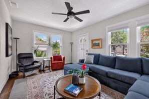 Un salon avec un canapé, une table basse sur laquelle repose un livre, un ventilateur de plafond et une vue par les fenêtres.