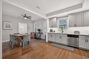 A modern kitchen with grey cabinets, a stainless steel dishwasher, and a dining area with a wooden table and grey chairs.