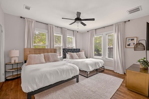 Une chambre avec deux lits, un ventilateur de plafond, de grandes fenêtres avec des rideaux, une table de chevet avec une lampe et une petite table d’appoint avec une plante.