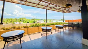 A covered outdoor seating area with tables and chairs, a view of greenery and distant buildings, and a clear sky.