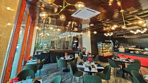 A restaurant interior with round tables, green chairs, hanging spherical light fixtures, and a kitchen area in the background.