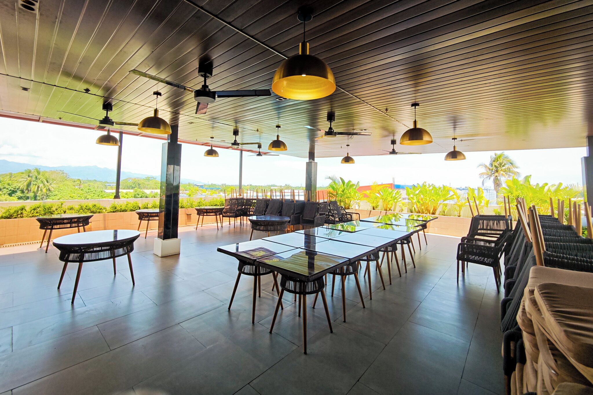 An outdoor dining area with tables and chairs, ceiling fans, and a view of greenery.