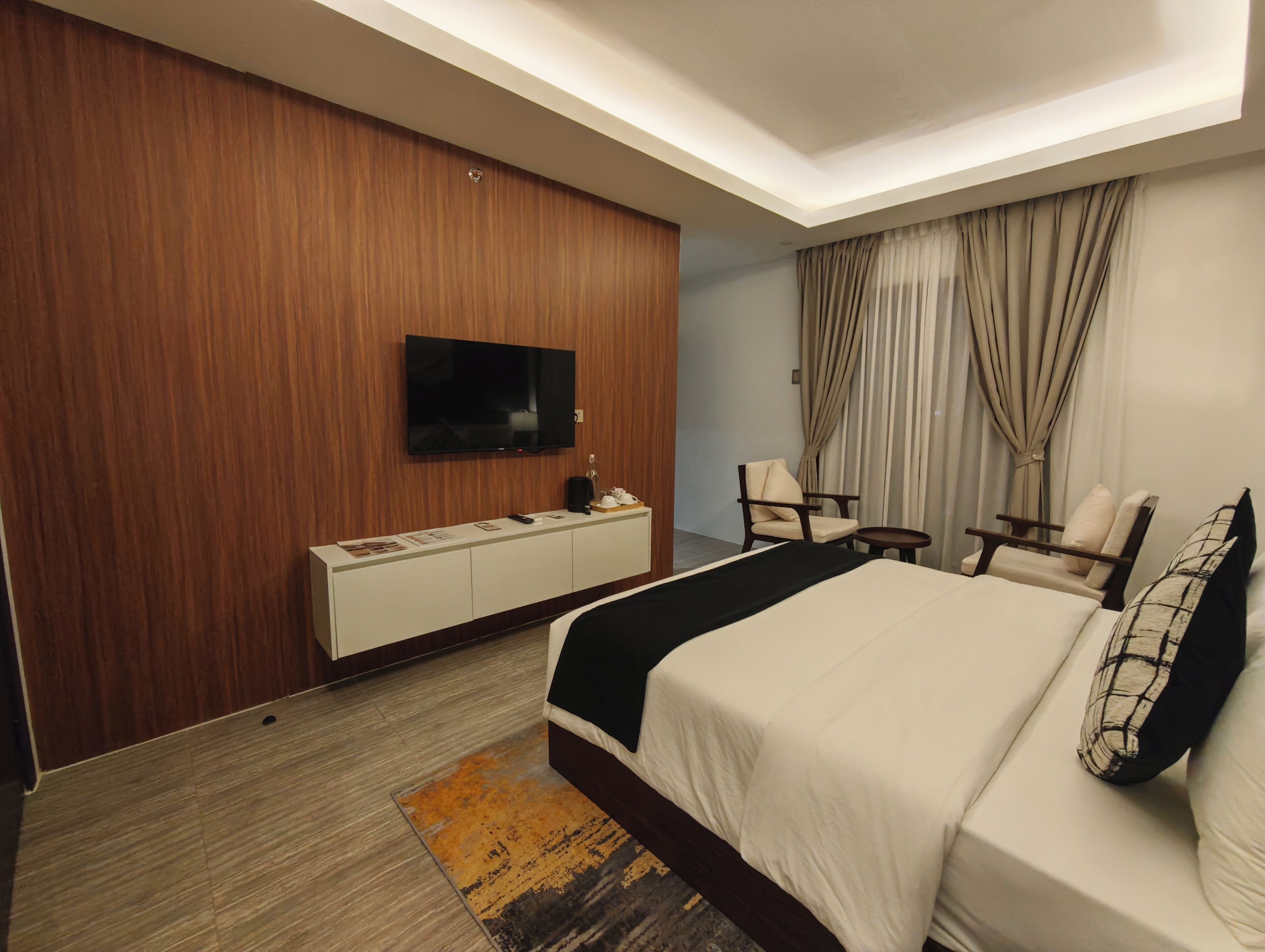 A hotel room with a bed, a TV mounted on the wall, a desk with a chair, and a window with curtains.