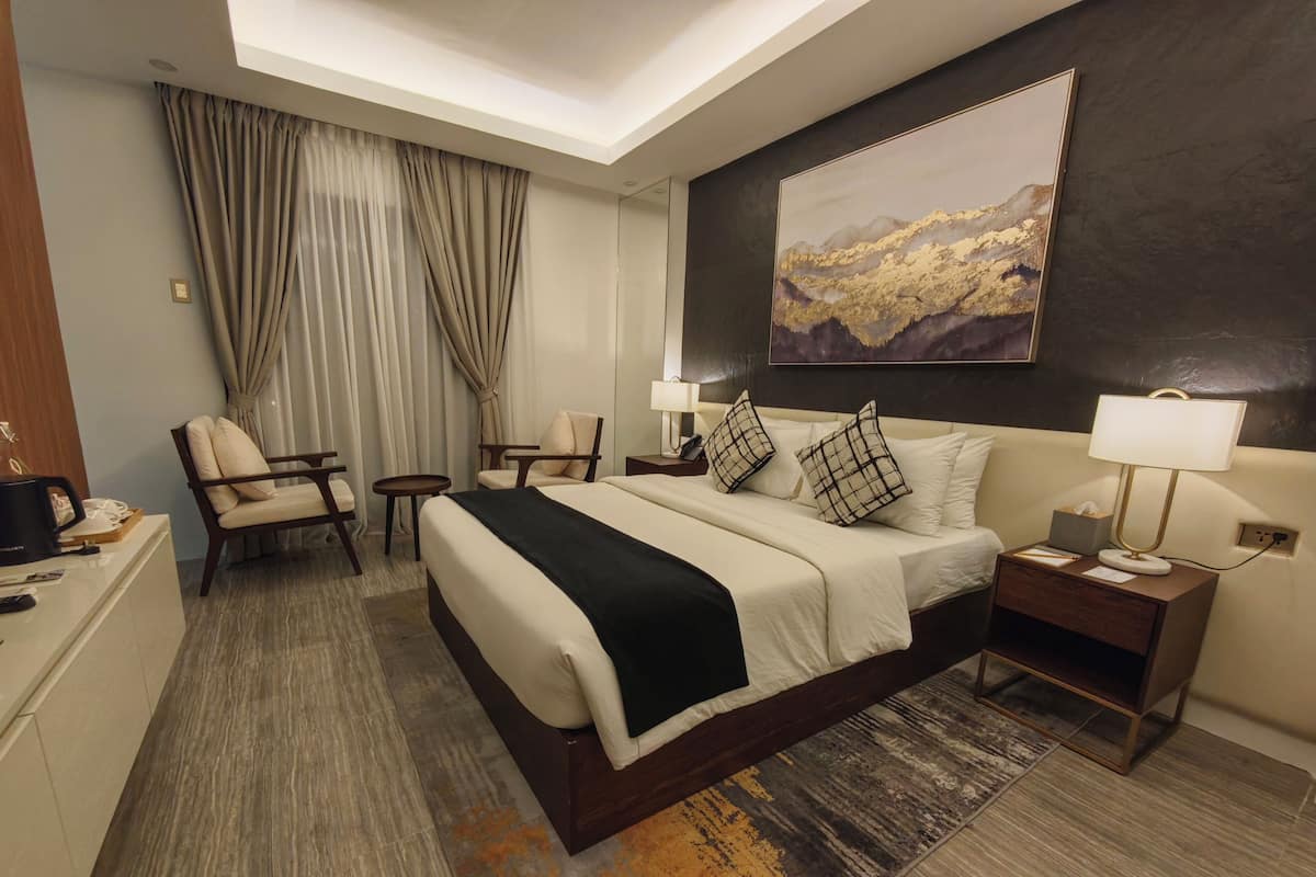 Una habitación de hotel con una cama grande, un escritorio, una silla, una mesita, una mesita de noche, una lámpara y un cuadro en la pared.