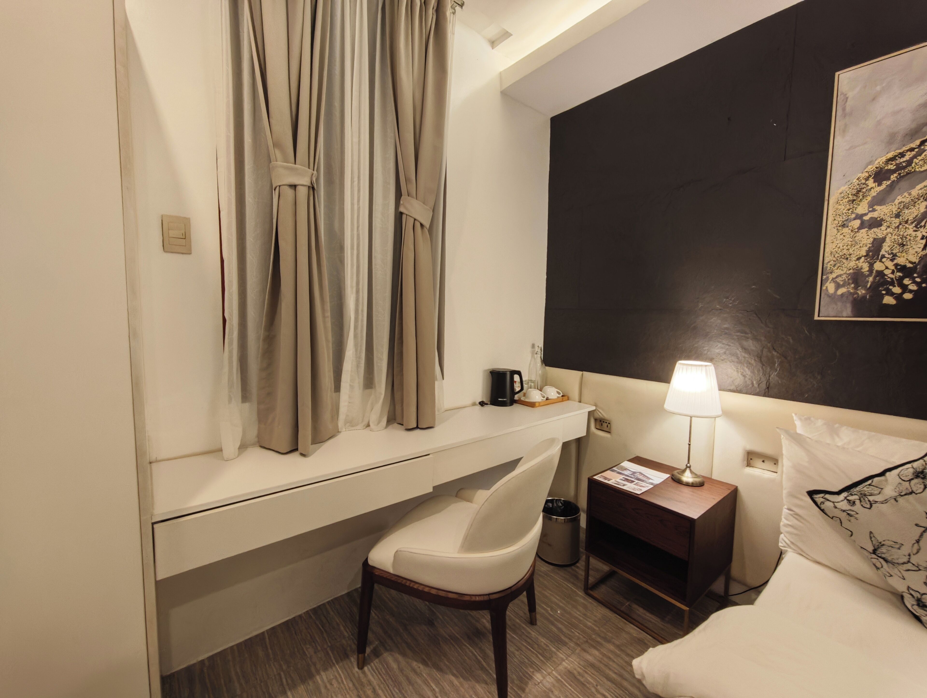 A hotel room with a bed, a chair, a bedside table with a lamp, a small table with a coffee maker, and a wall with a painting.