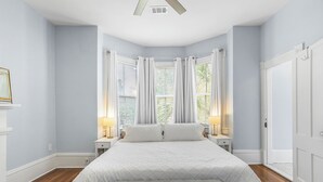 Un dormitorio con una cama grande, dos mesitas de noche, un banco y una ventana con cortinas.