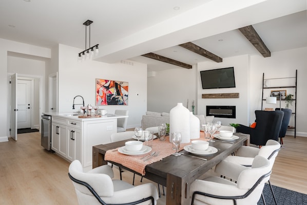 A modern dining area with a wooden dining table set for six, white chairs, and a kitchen in the background.