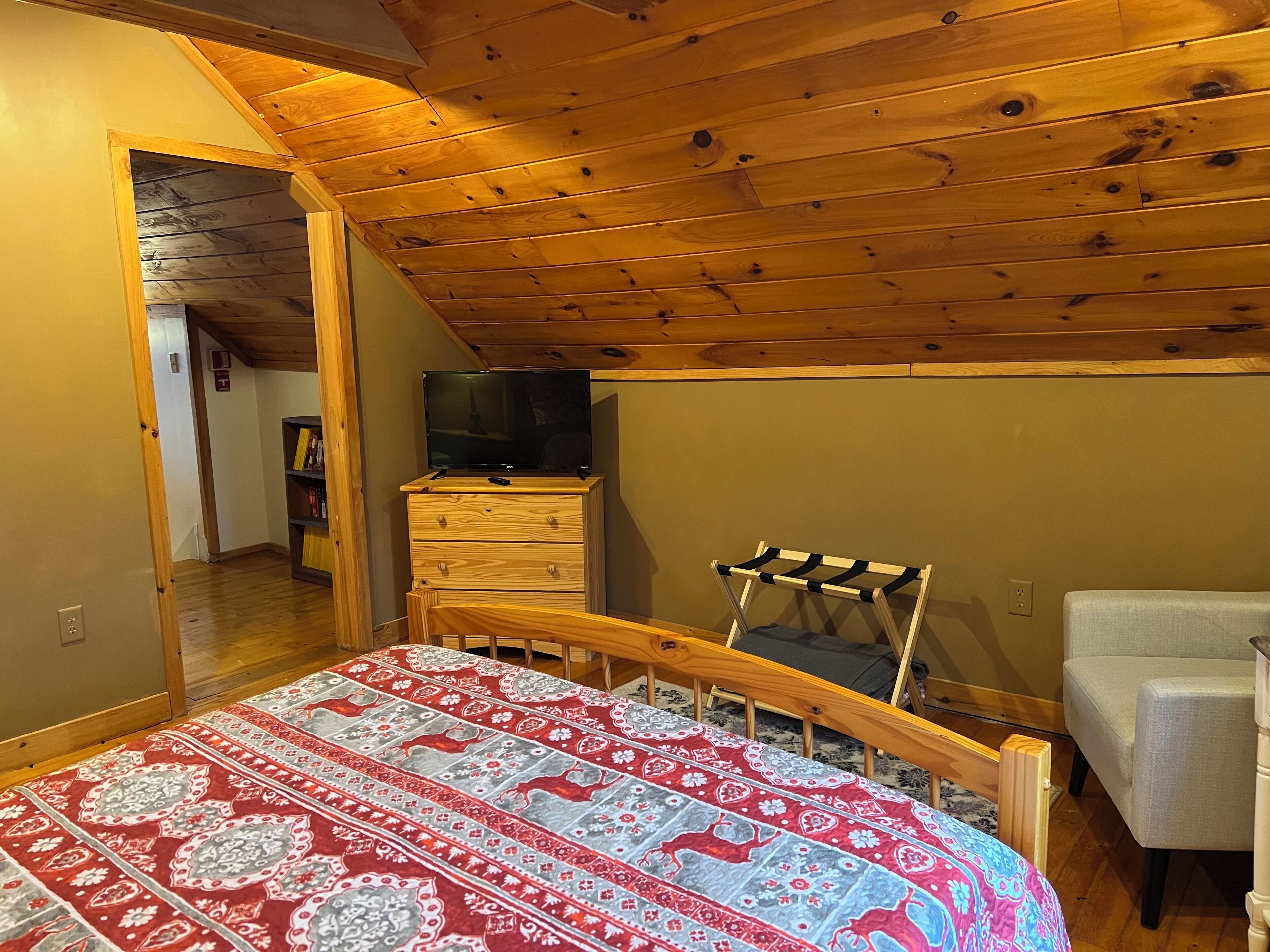 A bedroom with a wooden ceiling, a bed with a patterned bedspread, a TV on a wooden dresser, and a chair.