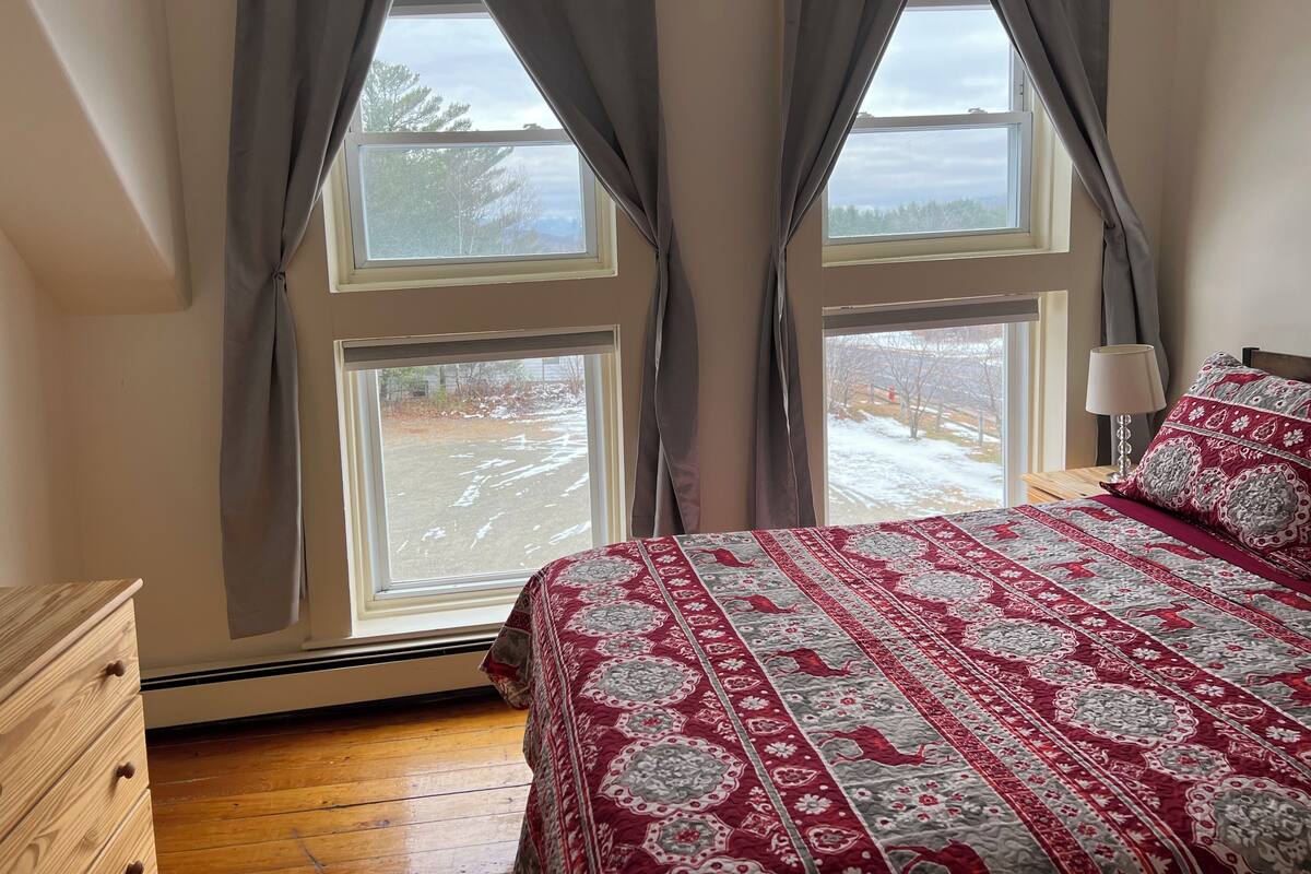 Une chambre avec deux fenêtres, un lit recouvert d’une couvre-lit à motifs, une commode en bois et une lampe sur une table de chevet.