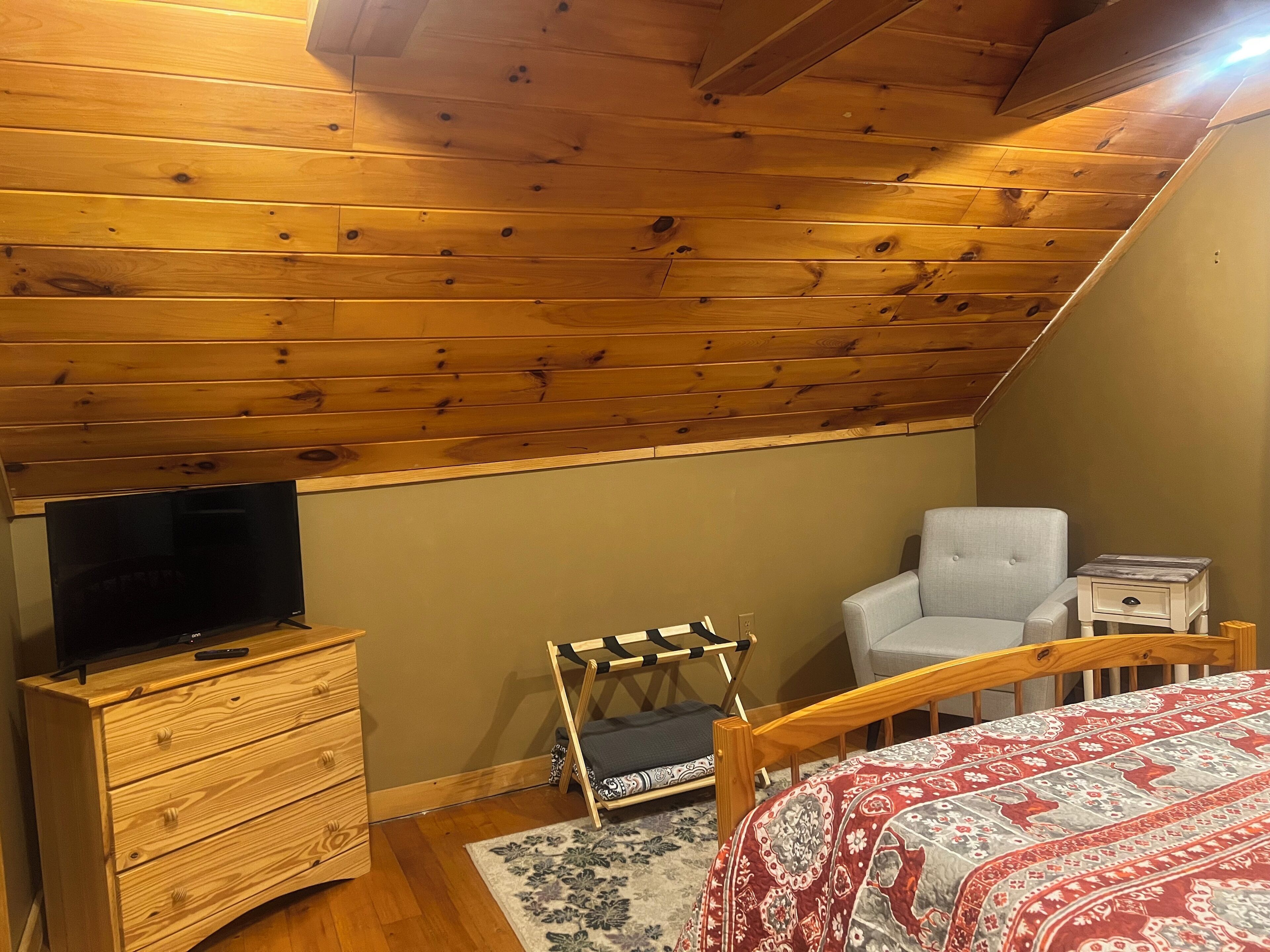 A bedroom with a wooden ceiling, a television on a wooden dresser, a white chair, a small bedside table, and a patterned bedspread.