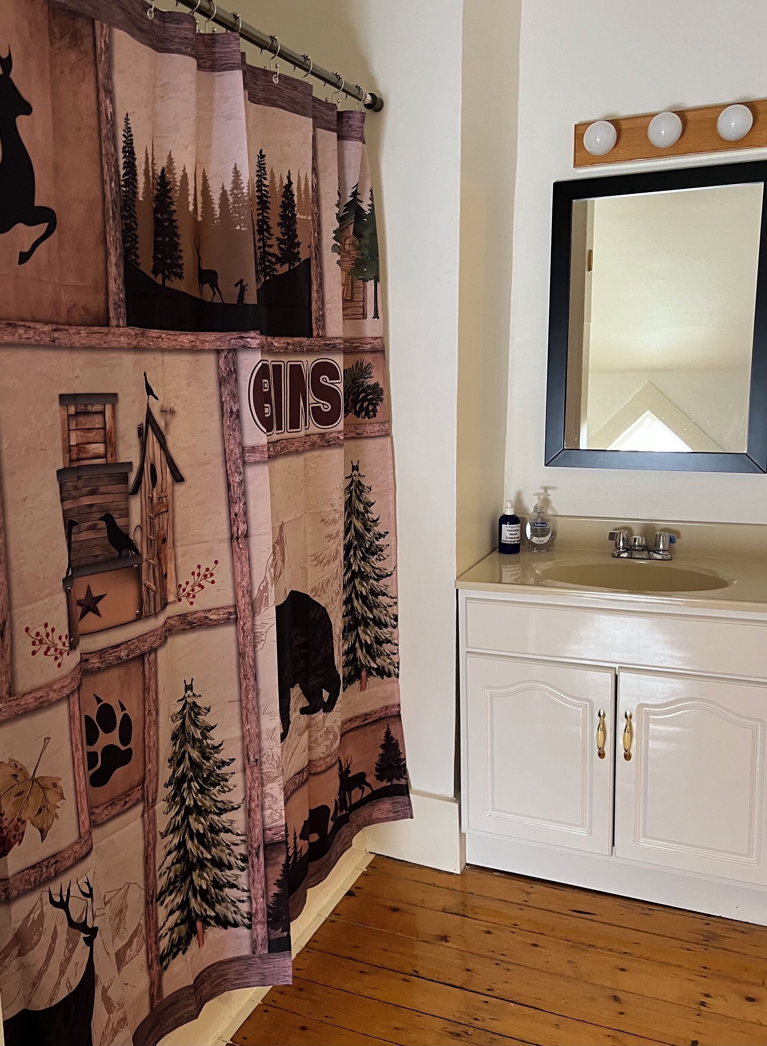 A bathroom with a wooden floor, a white sink with a mirror above it, and a shower curtain with a forest and animal print.