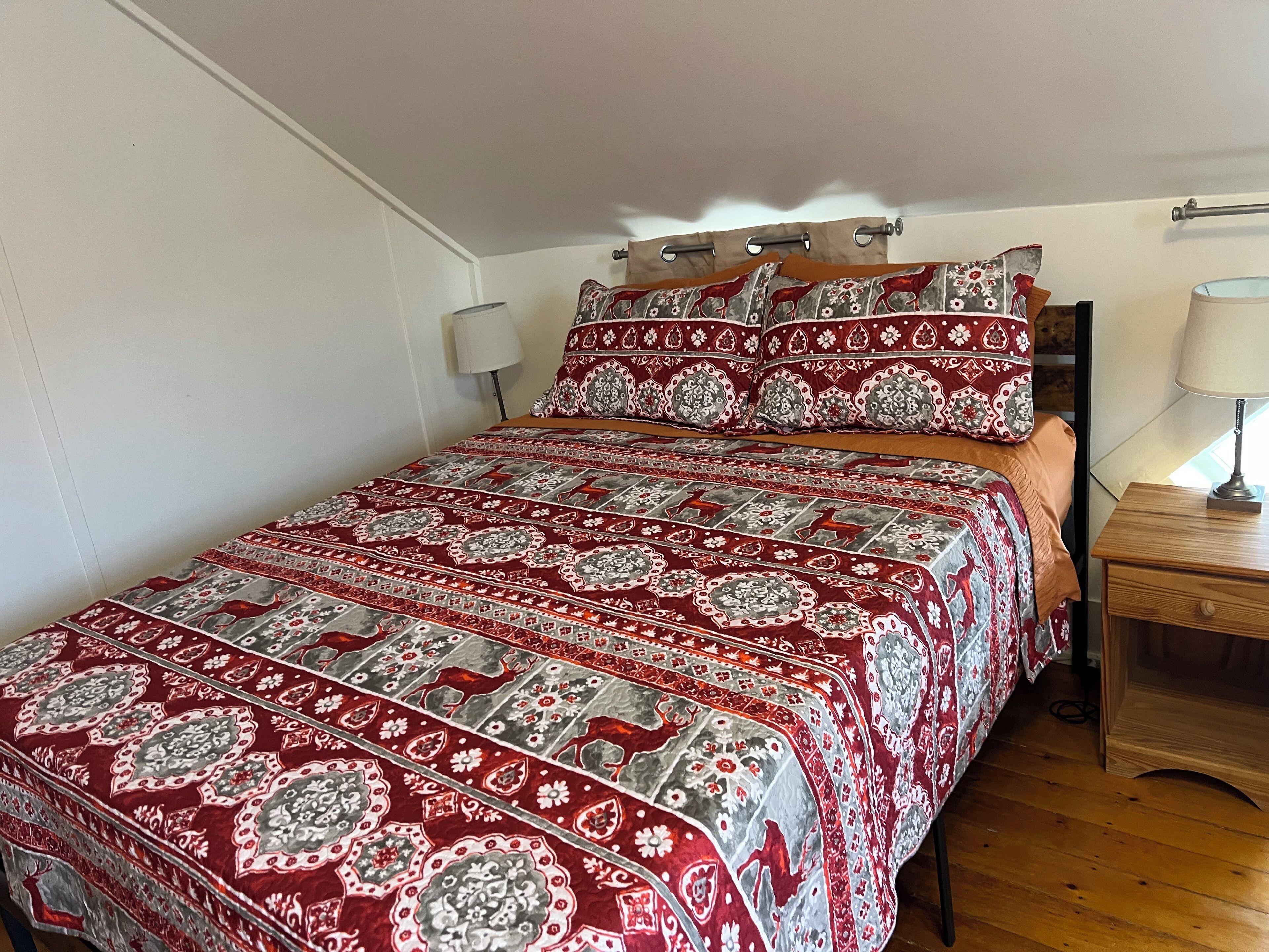 A bedroom with a bed covered in a patterned bedspread, a wooden nightstand with a lamp, and a window with curtains.