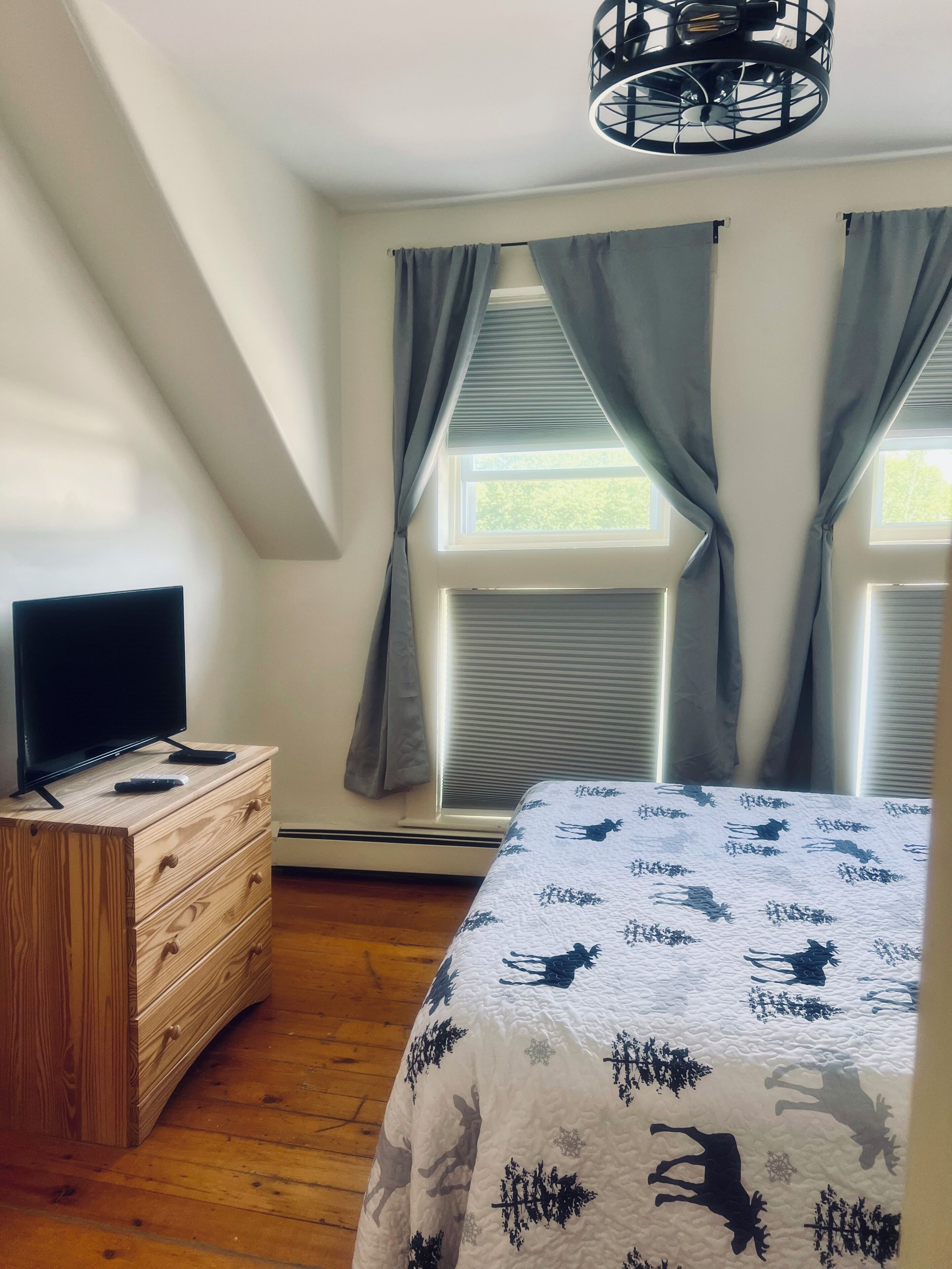 Une chambre à coucher avec un lit, une commode, un téléviseur posé sur la commode et une fenêtre avec des rideaux.