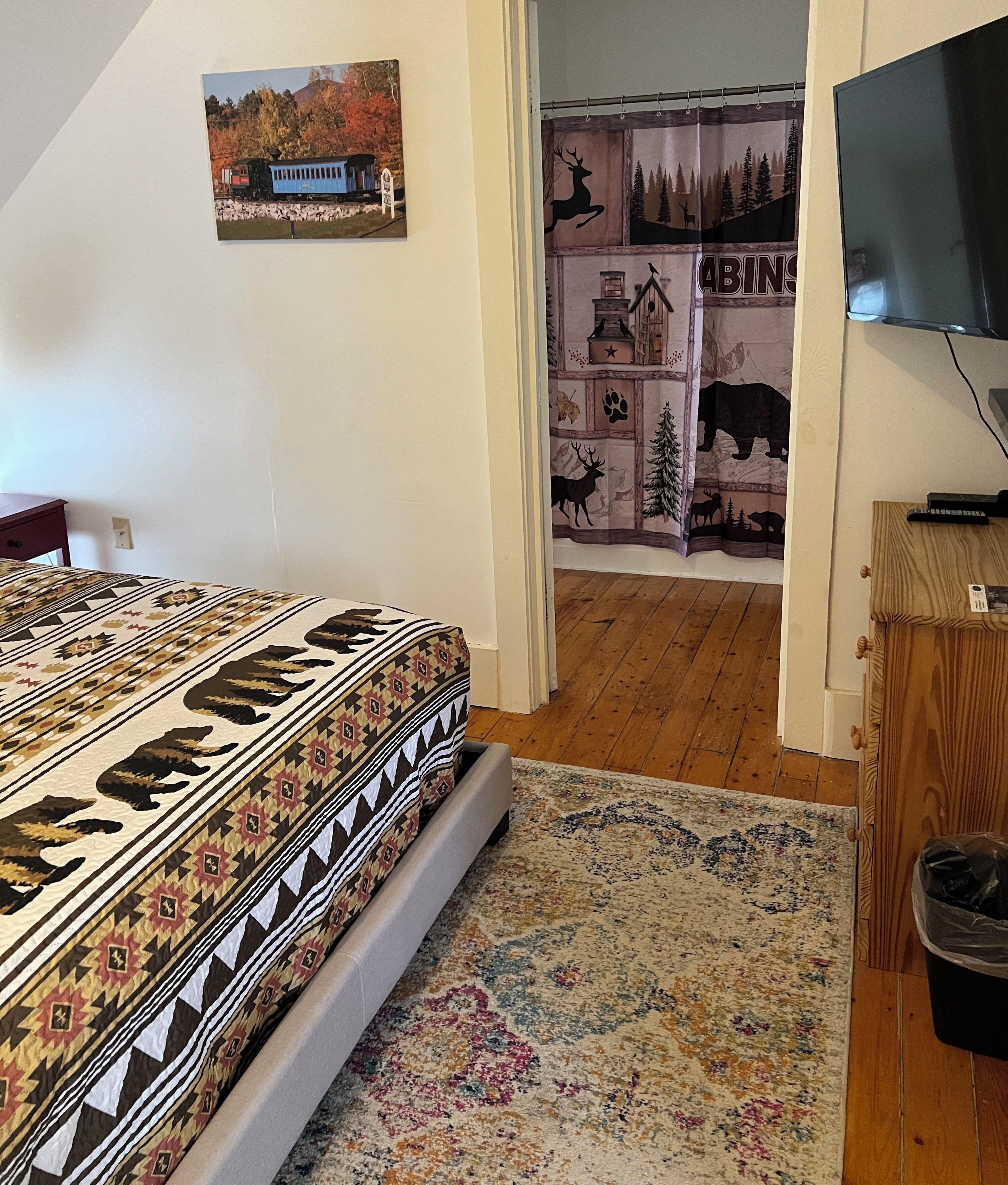 A bedroom with a bed, a TV, a wooden cabinet, and a wall with a forest-themed tapestry.