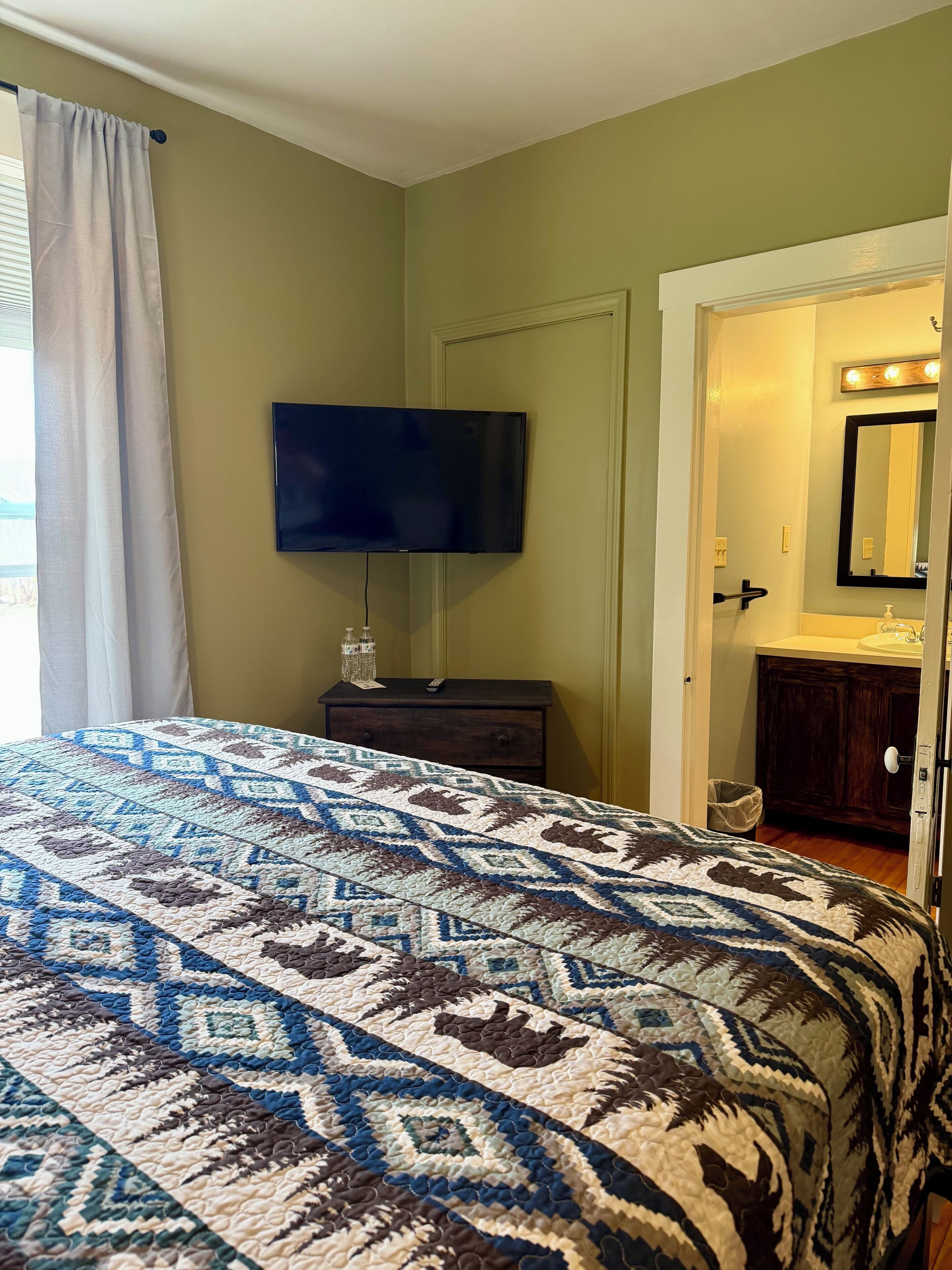 Un dormitorio con cama, televisor montado en la pared, cómoda y baño visible a través de una puerta abierta.