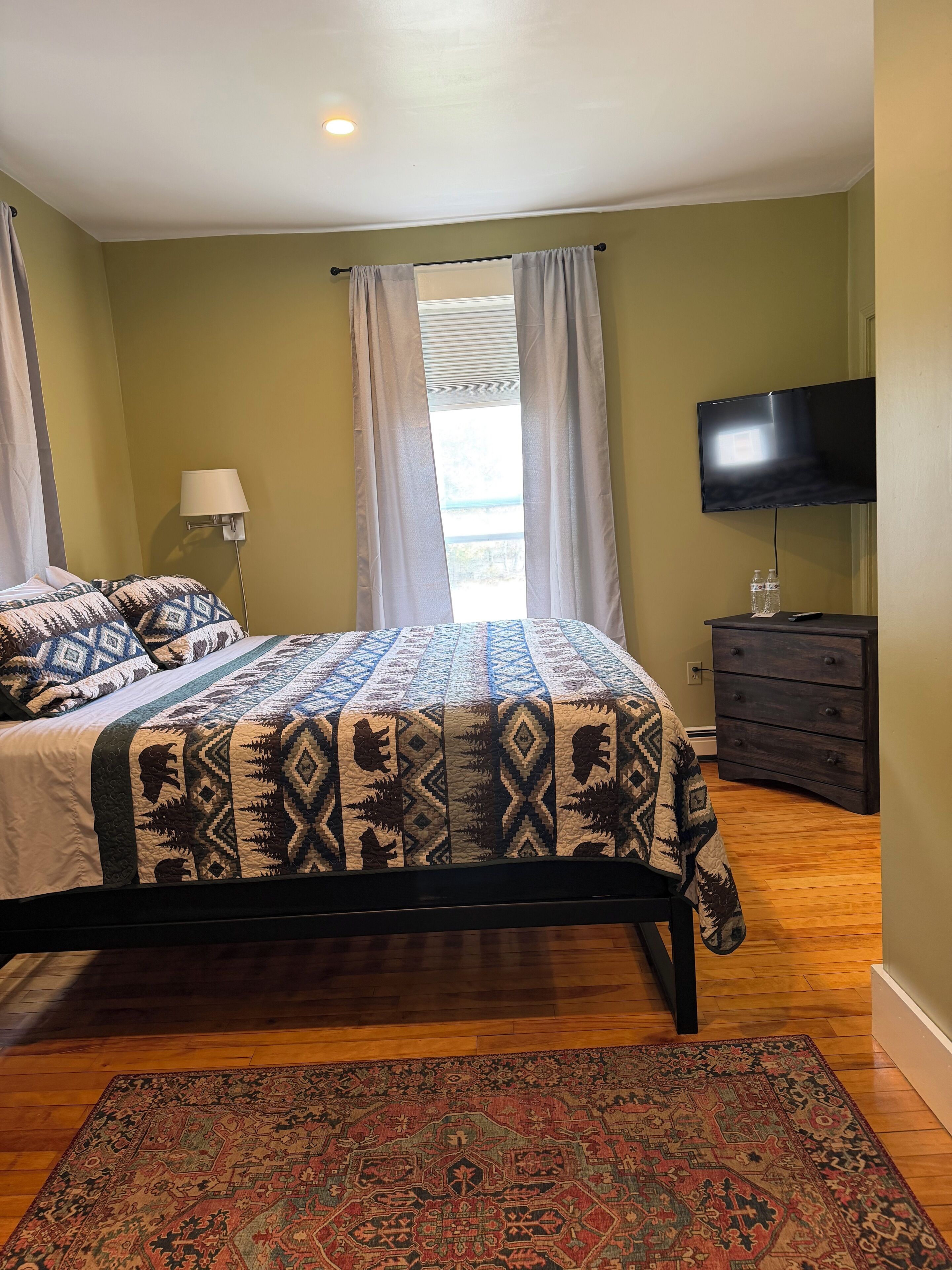 A bedroom with a bed, a TV, a dresser, and a window with curtains.