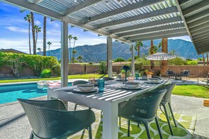 An outdoor patio with a table set for dining, surrounded by chairs, and overlooking a swimming pool and mountains.