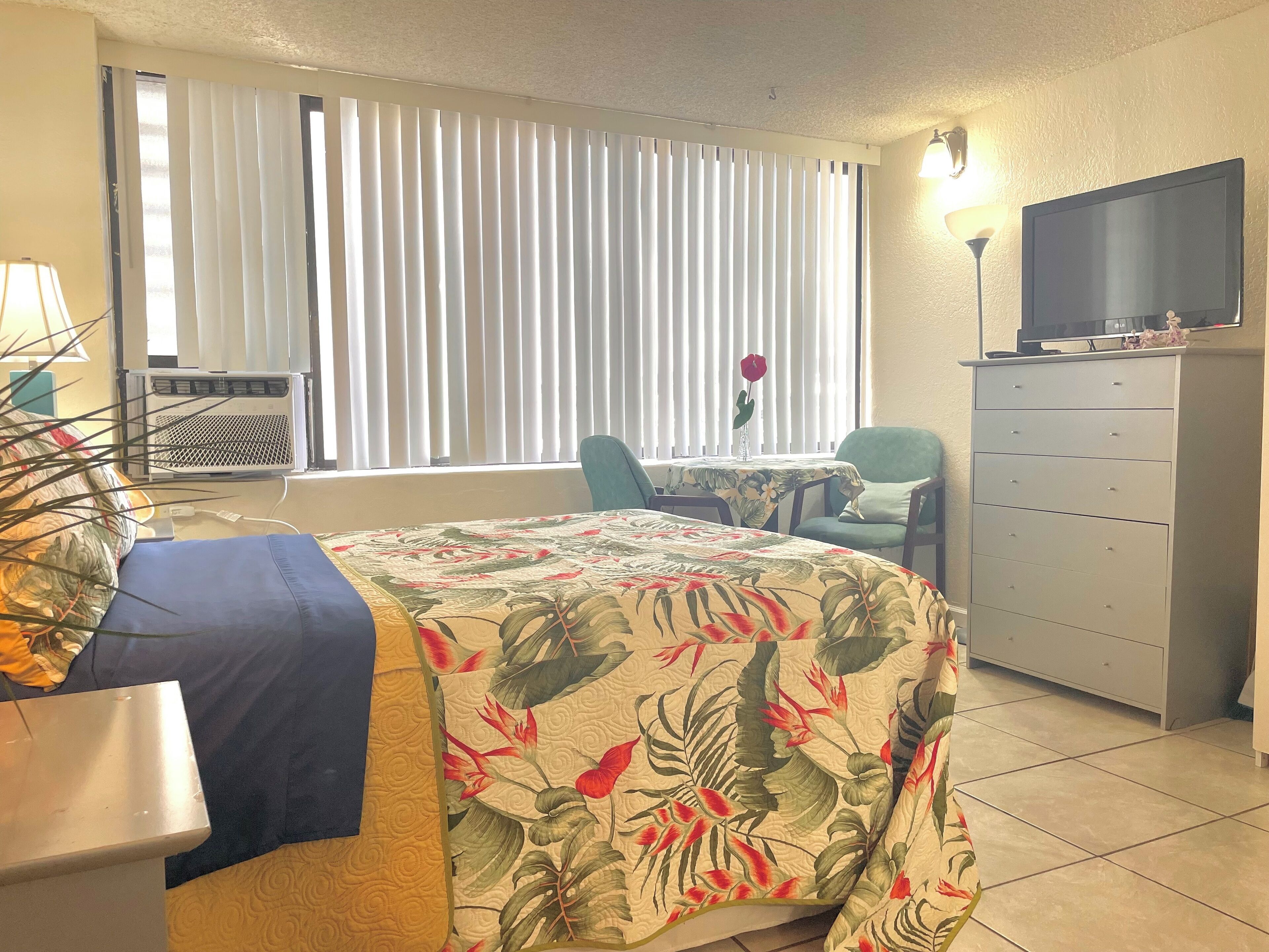 A hotel room with a bed, a desk, a chair, a dresser, a television, and a window with blinds.