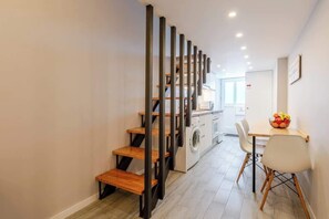 Una escalera con escalones de madera y barandillas negras que conduce a una zona de cocina con lavadora y una mesa con sillas.