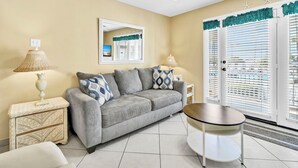 Un salon avec un canapé gris, deux tables d’appoint avec des lampes, un ventilateur de plafond et une vue à travers une porte équipée de stores.