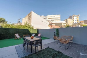 A rooftop patio with a wooden table and chairs, surrounded by a green wall and a view of city buildings.