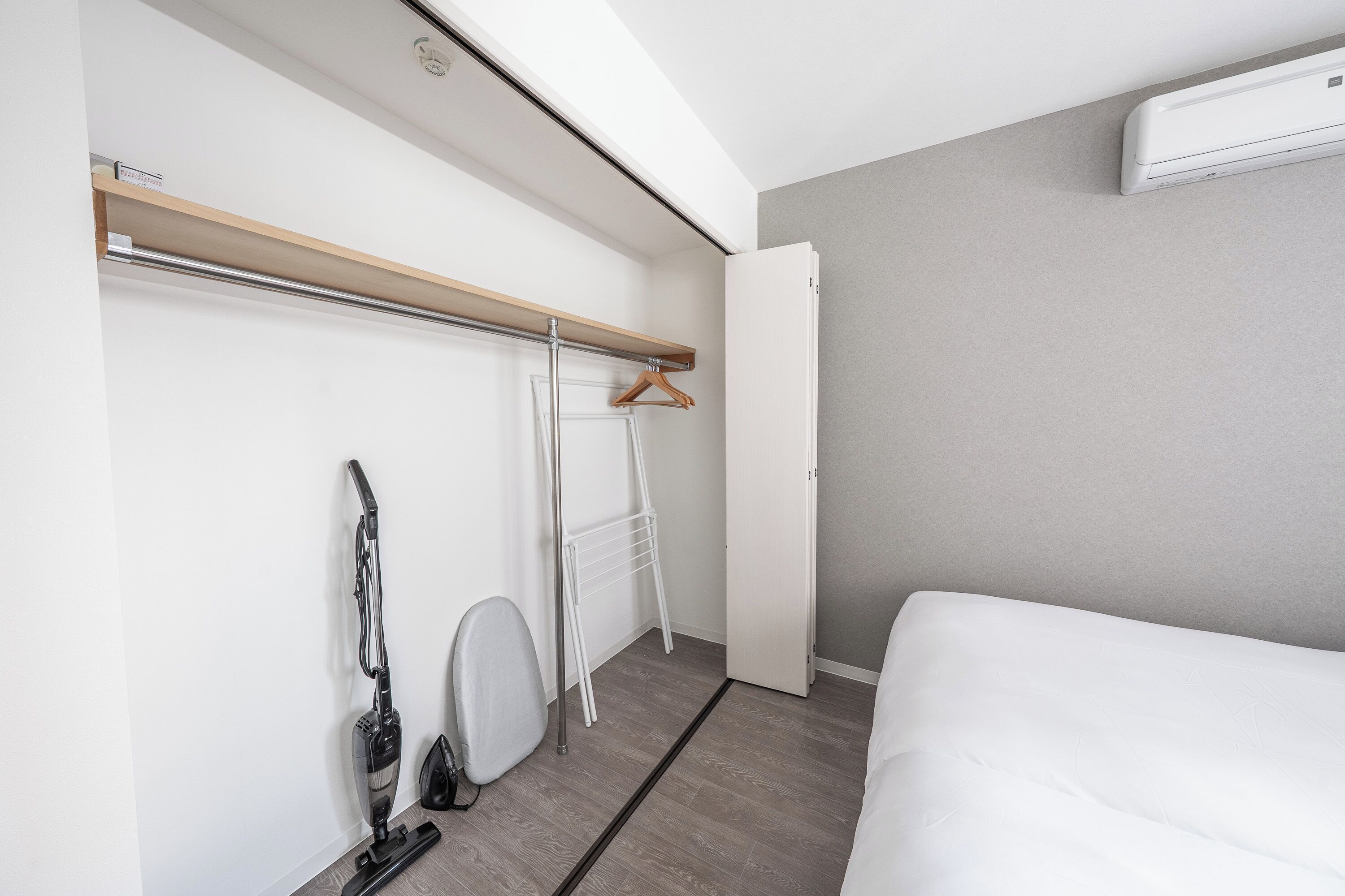 A bedroom with a bed, a vacuum cleaner, a drying rack, and a white wall with a window.