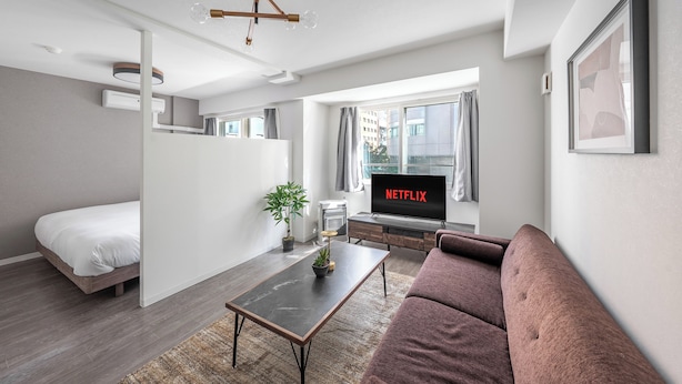 A modern living room with a sofa, a coffee table, a TV displaying 'NETFLIX', a bed, and a potted plant. - BELLE NEIGE 305 (Sapporo)