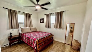 A bedroom with a wooden bed, a nightstand, a ceiling fan, and a window with curtains. - Homestead House - Simple comfort in the heart of Kansas (Hoisington)