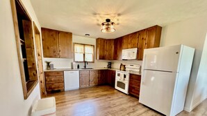 A kitchen with wooden cabinets, a white dishwasher, a white stove, a white refrigerator, and a window with curtains. - Homestead House - Simple comfort in the heart of Kansas (Hoisington)