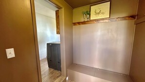 A narrow hallway with a wooden floor, a door on the left, and a wall-mounted shelf with two vases and a framed picture. - Homestead House - Simple comfort in the heart of Kansas (Hoisington)