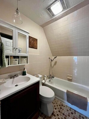 Une salle de bain avec un lavabo blanc, un meuble-lavabo, des toilettes et une baignoire. On trouve un porte-serviettes au-dessus de la baignoire et un élément décoratif sur le mur.