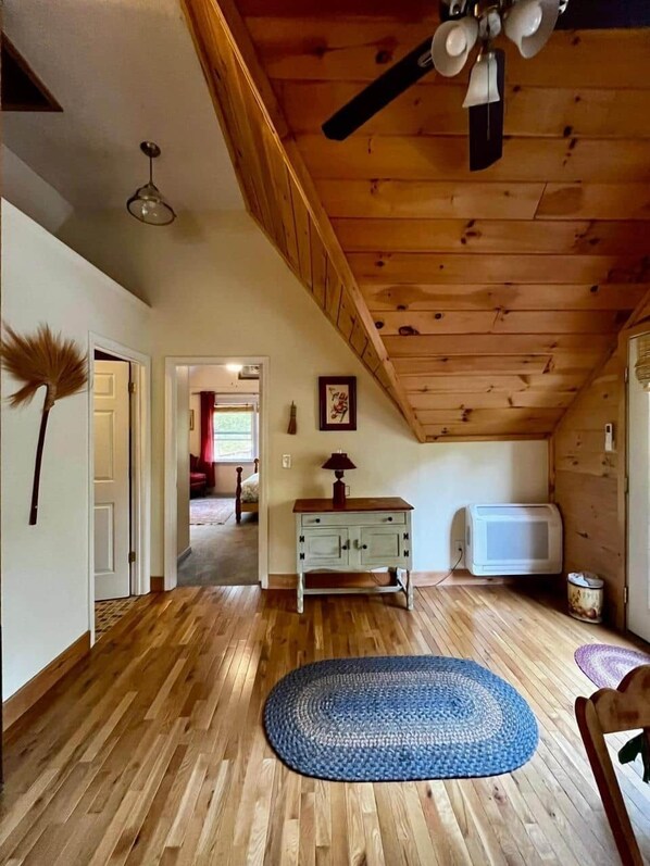 A room with wooden flooring, a ceiling fan, a white door, a blue rug, a wooden cabinet, and a microwave.