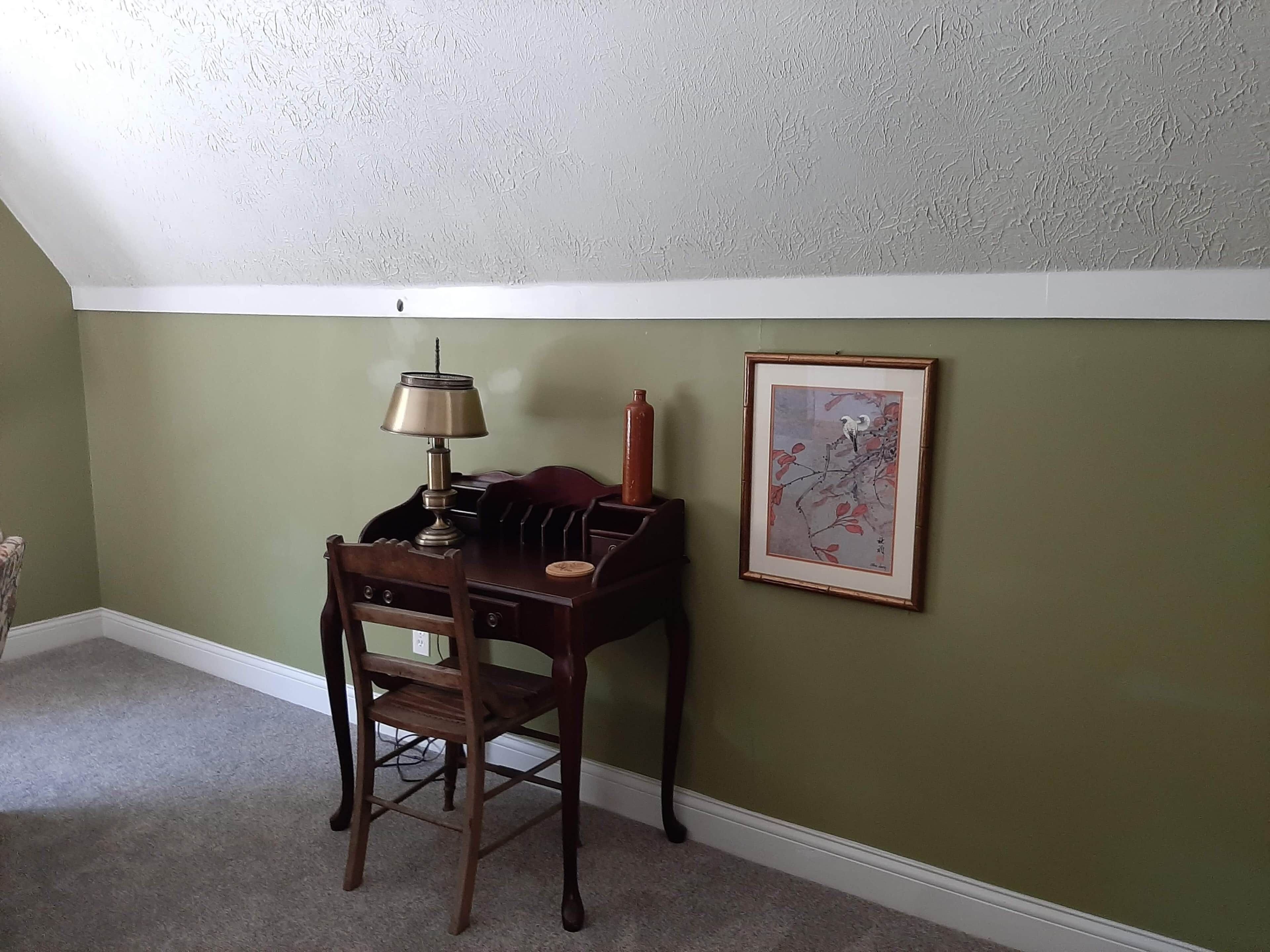 Une pièce d’angle avec un bureau en bois, une lampe, un tableau encadré et une chaise.