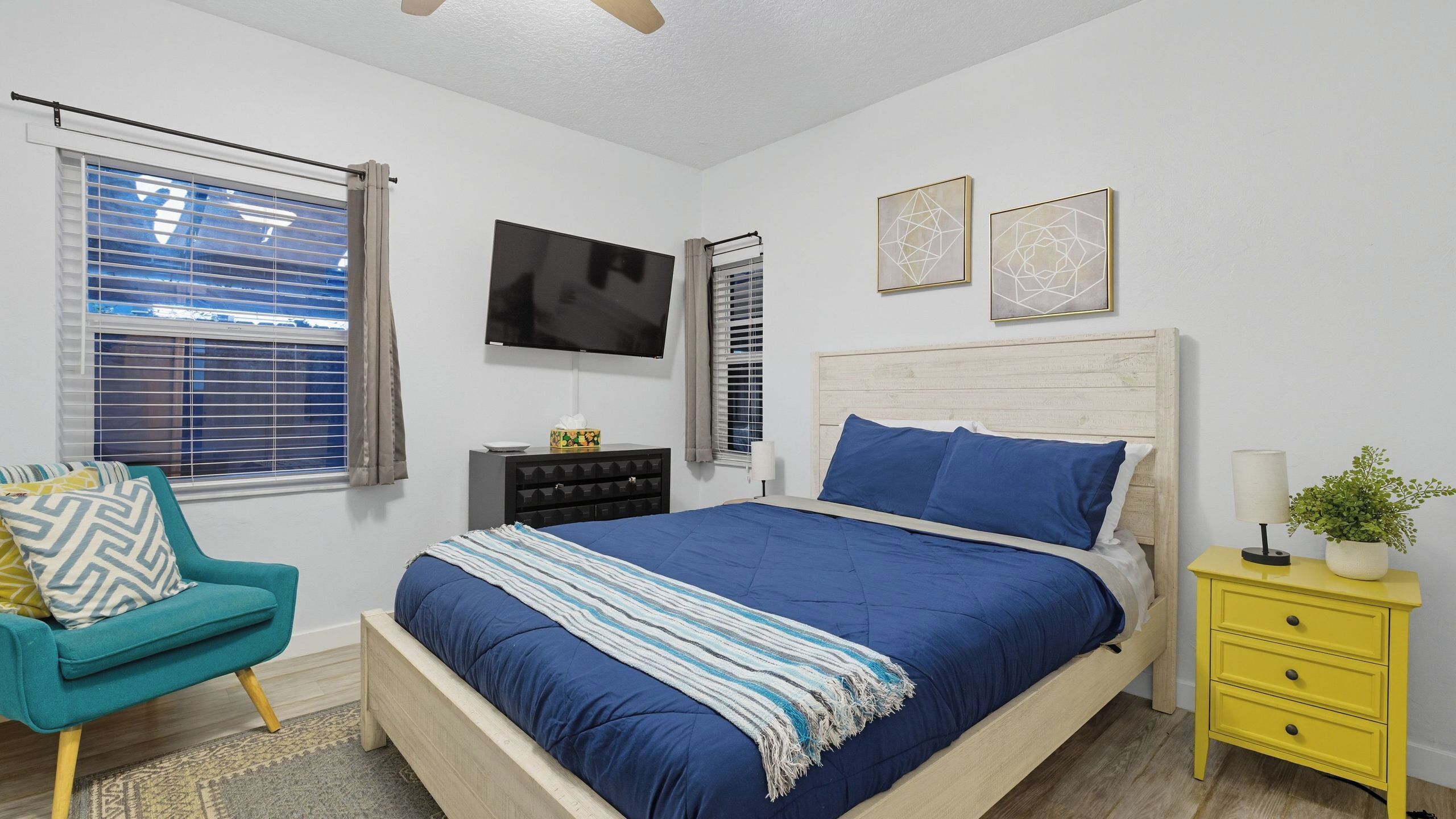 A bedroom with a bed, a TV, a chair, a bedside table with a lamp, and artwork on the wall.
