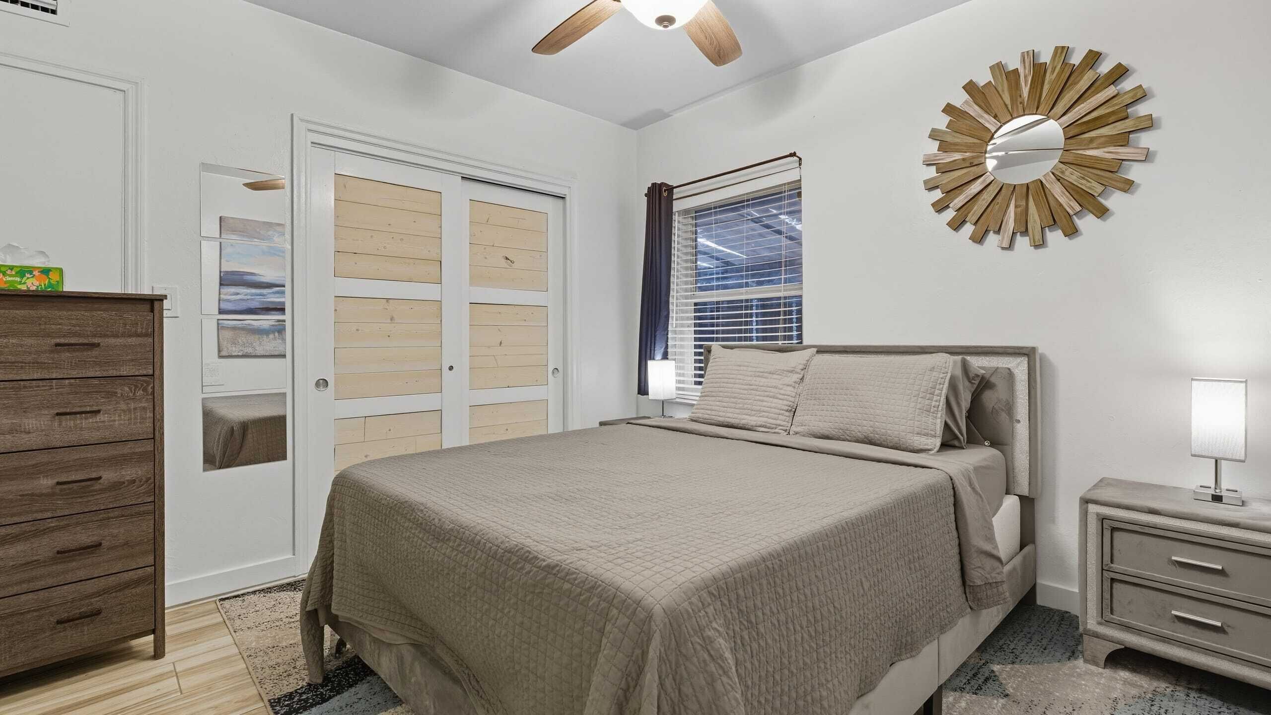 A bedroom with a bed, a nightstand, a dresser, a ceiling fan, and a window with blinds.