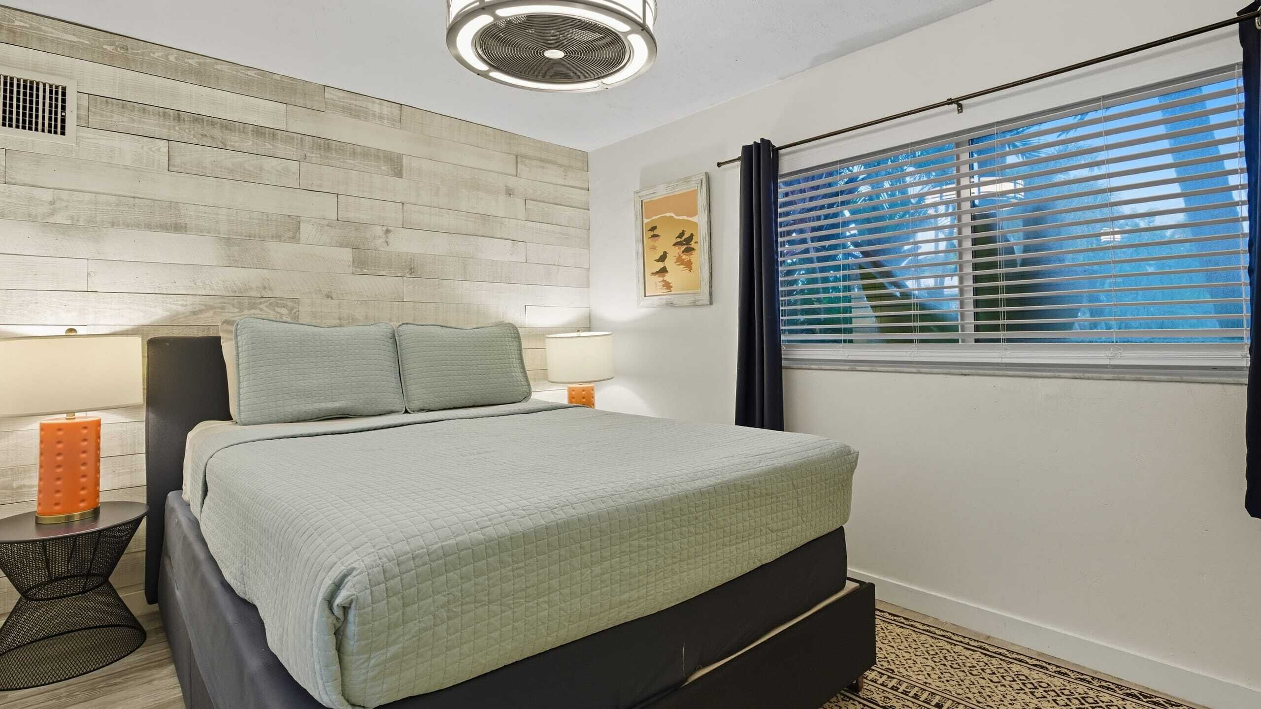 A bedroom with a bed, two bedside lamps, a wall-mounted heater, a window with blinds, and a patterned rug.